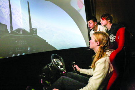 Wings Aerospace Academy students fly an aircraft in the AviationExtreme flight simulator.  Angelica Harvey is at the controls as Brandon Holleman (left rear) and Rocket Haverland stand beside her.