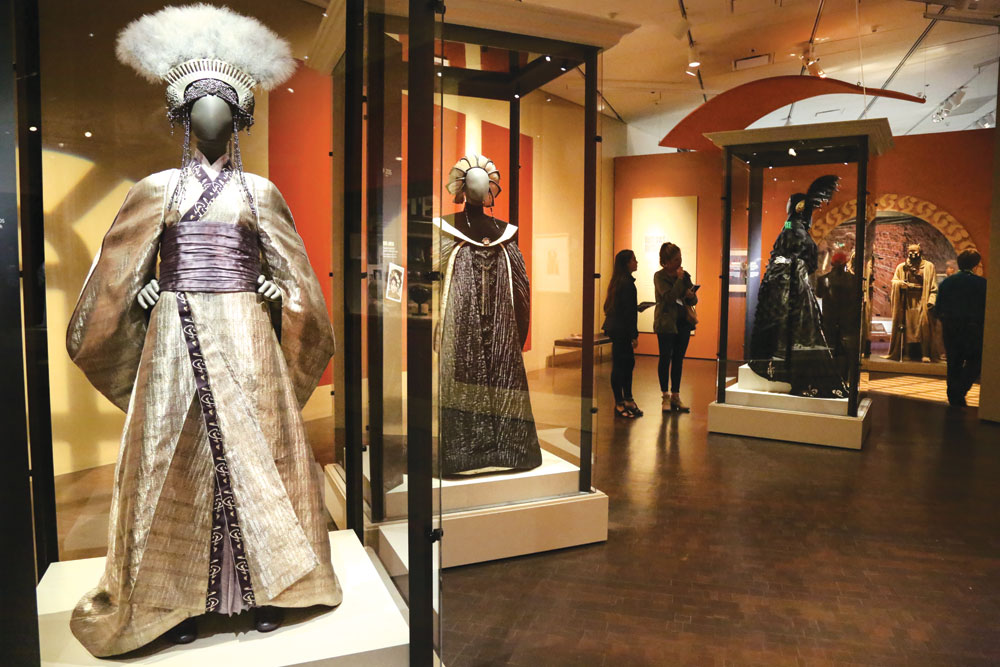 The exhibit explores the connections between costume and character with recognizable costumes in 13 thematic sections—including Jedi vs. Sith and the Galactic Senate. Focusing on the creative process of bringing characters to life, the exhibit features many original designs, as well as an art studio, costume shop and fitting room. In the foreground is the gown Queen Apailana wore at the funeral of Padme Amidala.